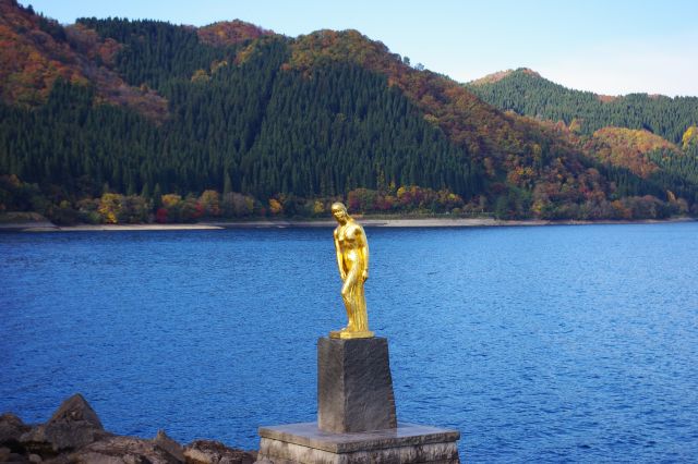 湖畔には黄金色の「たつこ像」が立っています。永遠の若さと美貌を願い湖神になったと伝えられています。奥には紅葉と深緑の針葉樹が混在する山。