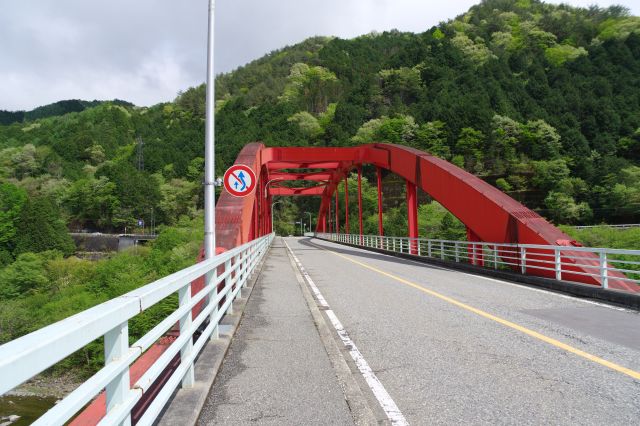 橋の歩道を進む。緑を背景とする赤い鉄橋が印象的です。